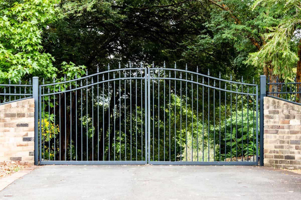 Metal driveway property entrance gates set in brick fence with garden trees in background

