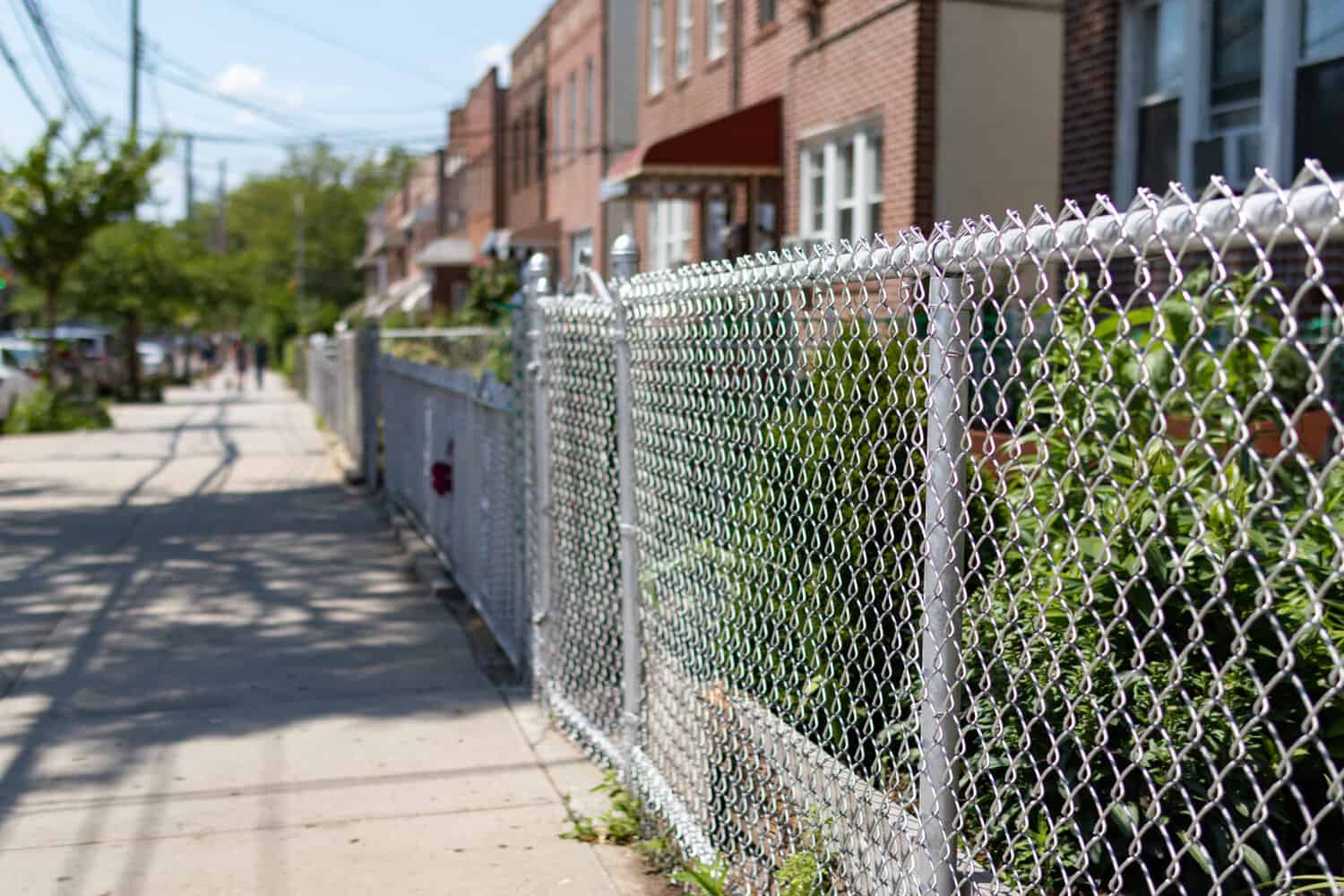 A chain link fence along an empty sidewalk with a row of old brick homes with gardens in Astoria Queens New York.