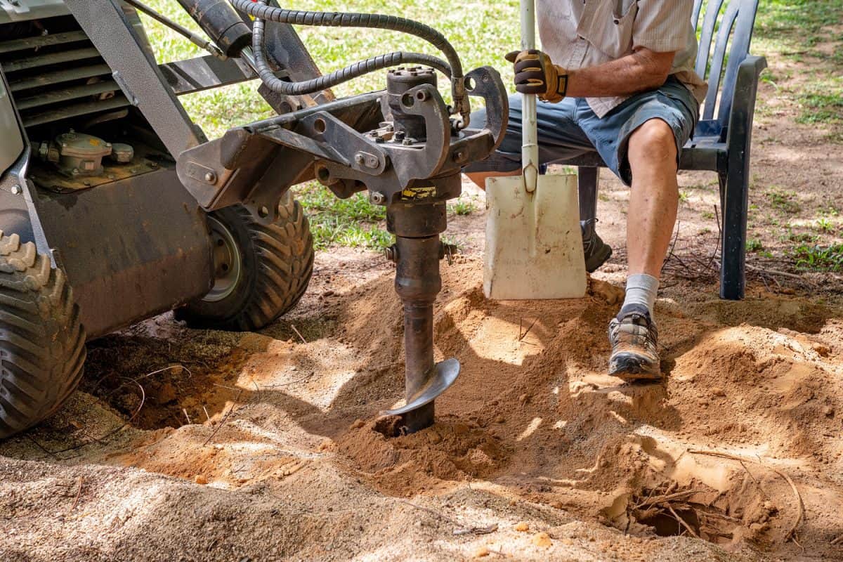 A mechanical auger digging holes in the soil with the aid of a senior male with a shovel

