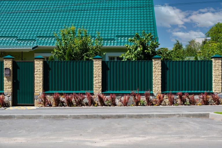 Green metal corrugated fence with brick pillars on the background of a beautiful house, Do Corrugated Metal Fences Rust?