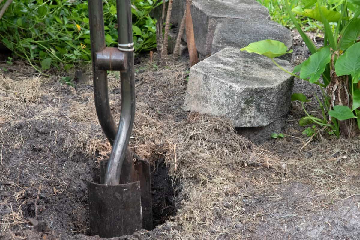 Steel post hole digger with shovel-like blades is being used to create a deep hole for a fence post with concrete bricks and green plants in the background.
