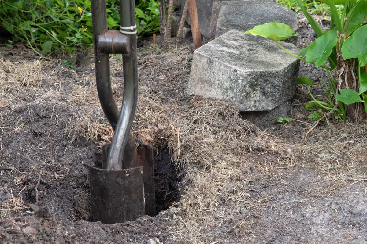 Steel post hole digger with shovel-like blades is being used to create a deep hole for a fence post with concrete bricks and gree