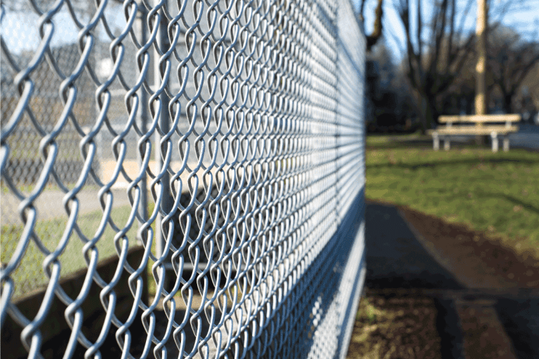 metal fence cage in a soccer field, park bench behind, sunny day, closeup, detail, horizontal. How Deep Should Chain Link Fence Posts Be Buried