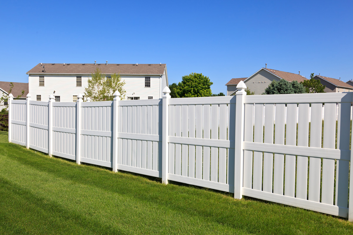 New and contemporary white vinyl fence in a nicely landscaped back yard with blue sky in the background.

