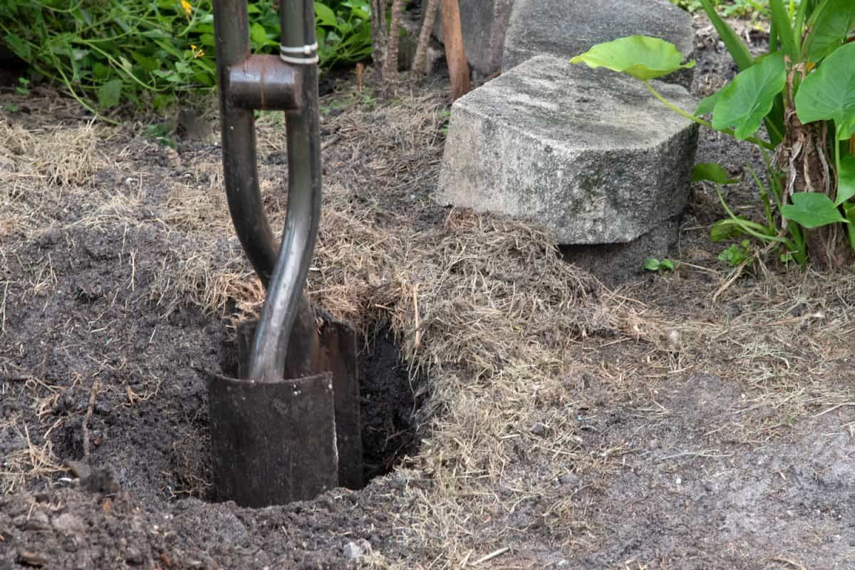 Steel post hole digger with shovel-like blades is being used to create a deep hole for a fence post with concrete bricks and green plants in the background.